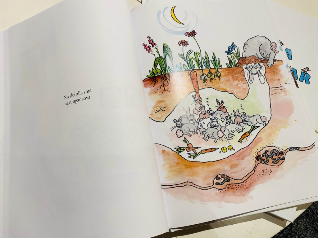 Uppslag som visar en illustration av en underjordisk kaninhåla med sovande småkaniner. Ovanpå marken syns blommor och växter. Texten lyder "Nu ska alla små harungar sova."